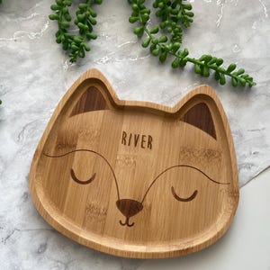 May include: A light brown wooden plate shaped like a fox with a cute face. The plate has the name "River" engraved on it.