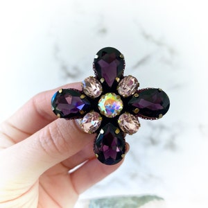May include: A purple and iridescent flower-shaped brooch with a gold setting. The brooch is made of rhinestones and has a cross-shaped design.