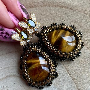 May include: A pair of gold earrings with a tiger's eye stone and a butterfly charm. The earrings are beaded with brown and black beads.