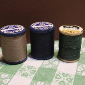 May include: Three spools of sewing thread. Two spools are blue with tan and dark blue thread, labeled Coats & Clark's Button & Carpet. The third spool is green thread with a label reading Talon, Color No. 788, and 15 cents. Each spool is cylindrical.