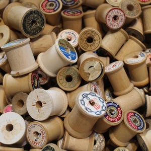 May include: A large collection of vintage wooden sewing thread spools piled together. Each spool features a cylindrical light wood body with a central hole and a circular paper label on the top end, displaying various brand names and text.