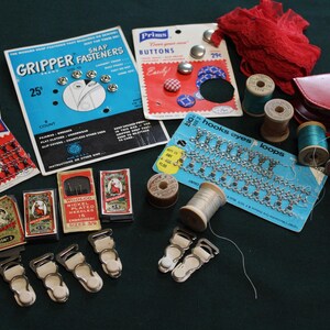 May include: A collection of vintage sewing notions including Prims brand snap fasteners, buttons, hooks and eyes, and nickel-plated needles in original packaging. Also includes wooden spools of thread, metal garter clips, red lace trim, and a red vinyl case.