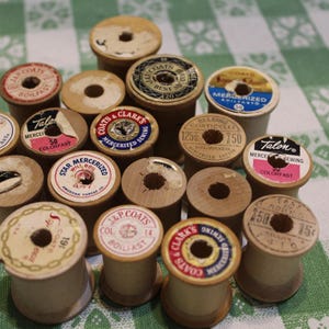 May include: A collection of vintage wooden sewing thread spools arranged in a cluster. Each spool features a unique circular paper label on the top, displaying various brand names like J. & P. Coats, Talon, and Belding Corticelli with text.