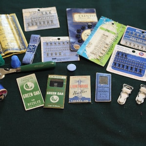 May include: A collection of vintage sewing notions including wooden spools of thread, metal thimbles, a tracing wheel, and various paper packets of needles, hooks, and eyes. Also included are several metal garment clips and a small measuring tape.