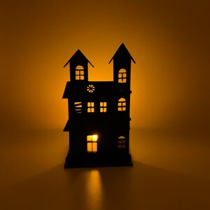 May include: A black metal candle holder in the shape of a haunted house with lit windows. The silhouette features two turrets and multiple windows, with a warm glow. A decorative item for Halloween.