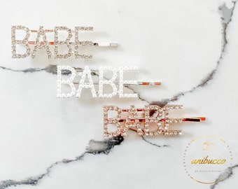 May include: Three silver hair clips with the word "BABE" spelled out in rhinestones. The clips are on a white marble background.