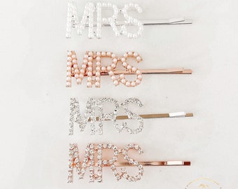May include: Four silver and gold hair clips with the word "MRS" spelled out in rhinestones and pearls. The clips are arranged vertically on a white background.