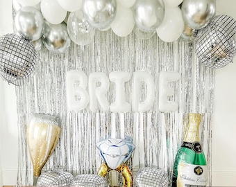 Op de afbeelding: Zilveren en witte ballon-slinger met het woord "BRIDE" in witte letters, een champagnefluit-ballon, een diamanten ring-ballon, een champagnefles-ballon en zilveren discobal-ballons. De slinger hangt voor een zilveren tinsel achtergrond.