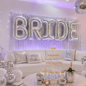 Puede incluir: Un sofá blanco decorado con globos de aluminio plateados que dicen BRIDE, un cojín BRIDE TRIBE y una torre de champán. La habitación cuenta con cortinas de flecos plateadas, bolas de discoteca, globos blancos y plateados y una caja de luz.