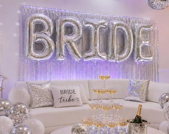 以下が含まれることがあります： BRIDEの文字の銀色のホイルバルーン、BRIDE TRIBEのクッション、シャンパンタワーで飾られた白いソファ。部屋には銀色のティンセルカーテン、ミラーボール、白と銀のバルーン、IN MY BRIDE ERAと書かれたライトボックスが配置されています。