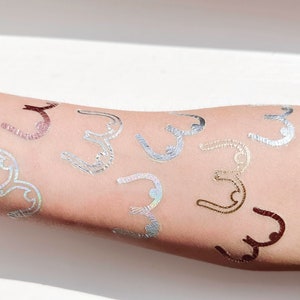 May include: A person's arm with multiple gold, silver, and iridescent temporary tattoos in the shape of a stylized letter "C".
