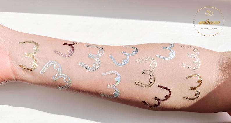 May include: A person's arm with multiple gold, silver, and iridescent temporary tattoos in the shape of a stylized letter "C".