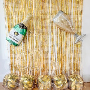 May include: A gold metallic fringe curtain backdrop featuring a large green champagne bottle balloon and a champagne flute balloon, with five gold square disco ball balloons arranged in a row along the bottom of the display.