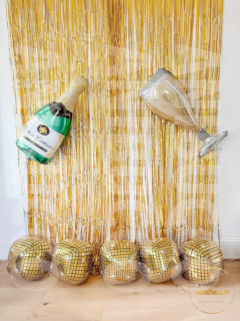 May include: A gold metallic fringe curtain backdrop featuring a large green champagne bottle balloon and a champagne flute balloon, with five gold square disco ball balloons arranged in a row along the bottom of the display.