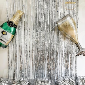 May include: A silver metallic fringe curtain backdrop featuring a foil balloon shaped like a green champagne bottle and a foil balloon shaped like a champagne flute, accompanied by five square silver disco ball balloons at the base.