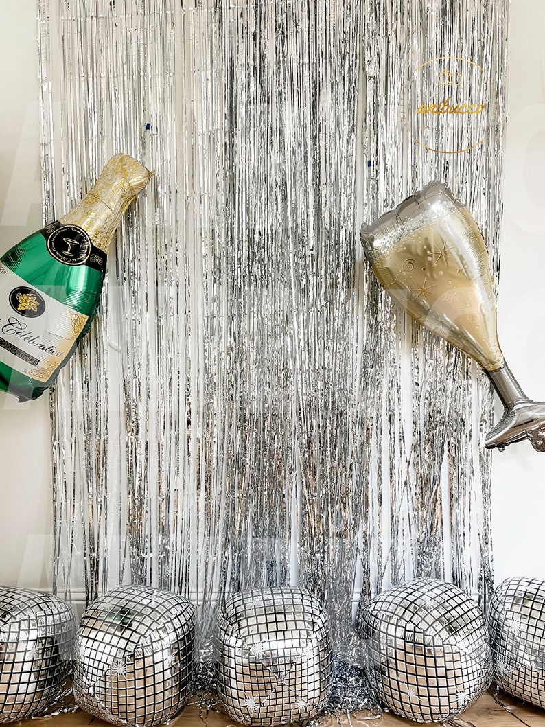 May include: A silver metallic fringe curtain backdrop featuring a foil balloon shaped like a green champagne bottle and a foil balloon shaped like a champagne flute, accompanied by five square silver disco ball balloons at the base.