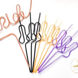 May include: A set of colorful reusable drinking straws shaped into different designs. The straws are pink, black, gold, and purple. The straws are bent into the shape of the word "bride" and the peace sign.