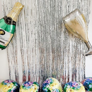 May include: A silver metallic fringe curtain backdrop featuring a green champagne bottle balloon, a gold champagne flute balloon, and five round disco ball balloons with multicolored square patterns and white starburst accents.