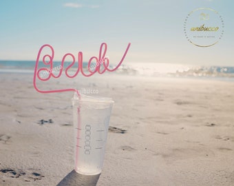 May include: A pink plastic straw bent into the word "beach" on a sandy beach with a clear plastic cup with a measurement scale on the side.