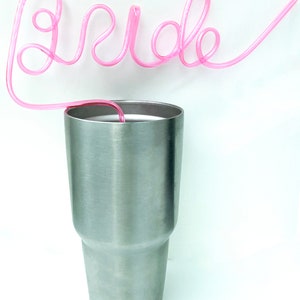 May include: A silver stainless steel tumbler with a pink plastic straw shaped to spell out the word "bride".