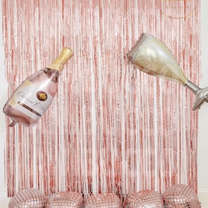 May include: A rose gold metallic fringe curtain backdrop featuring a champagne bottle balloon and a champagne flute balloon. Five square disco ball balloons are arranged in a row at the base. The bottle label reads Chateau Celebrate.