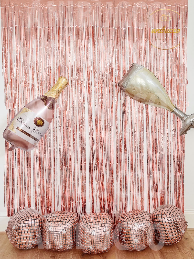May include: A rose gold metallic fringe curtain backdrop featuring a champagne bottle balloon and a champagne flute balloon. Five square disco ball balloons are arranged in a row at the base. The bottle label reads Chateau Celebrate.