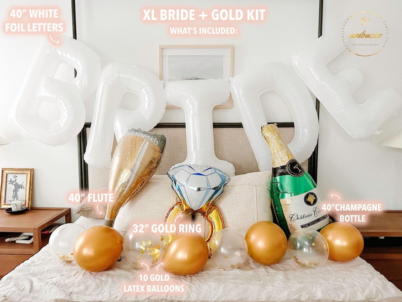 Extra large white letters spelling BRIDE hang above the bed in an arch. Below the letters on the bed rests three large complementing foil balloons, a large flute, a gold ring, a green champagne bottle and 10 gold chrome and confetti filled balloons