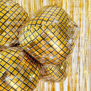 May include: A cluster of round, gold-colored foil balloons featuring a reflective, grid-like pattern that mimics a disco ball. Each balloon surface is decorated with small white starburst accents, set against a backdrop of shimmering gold fringe.
