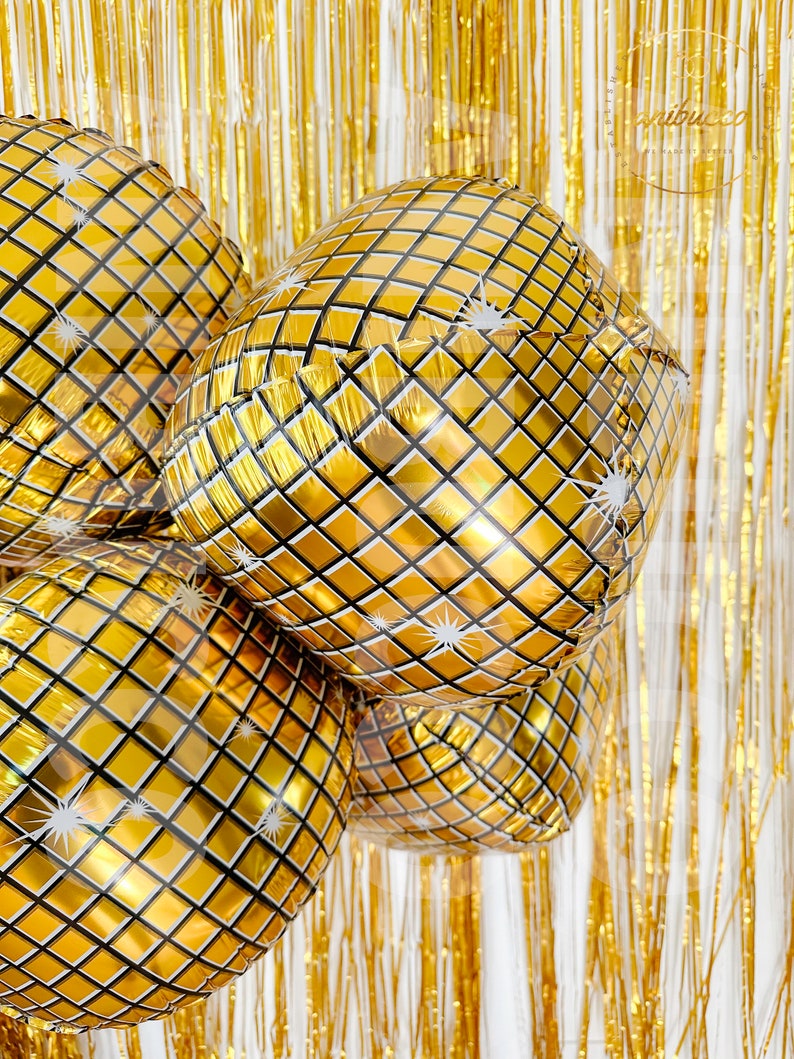 May include: A cluster of round, gold-colored foil balloons featuring a reflective, grid-like pattern that mimics a disco ball. Each balloon surface is decorated with small white starburst accents, set against a backdrop of shimmering gold fringe.