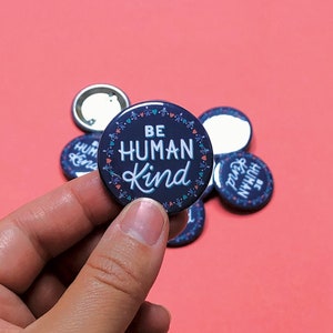 May include: A close-up of a hand holding a blue button with the text "Be Human Kind" in white lettering. The button is surrounded by a white border with small hearts and dots.