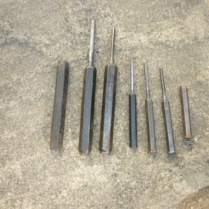 May include: A selection of metal punches, each with a dark, hexagonal handle and a pointed tip, laid out on a concrete surface. The tools vary in length, with measurements in centimetres, suitable for different applications.
