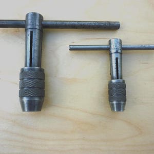 May include: Two metal tap wrenches, one large and one small, with textured handles and T-shaped handles. The tools are made of a dark, possibly steel, material and are designed for use with taps. They are set against a light wood background.