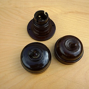 May include: Three dark brown, glossy electrical components consisting of one circular light bulb socket with a mounting flange and two round, dome-shaped light switches, each featuring a small central toggle lever on the top surface.