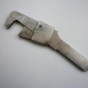 May include: A silver-coloured vintage adjustable wrench. The tool has a curved head and a sliding jaw, with a long handle. The wrench shows signs of wear and age, with a slightly tarnished appearance. The tool is made of metal.