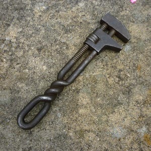 May include: A dark metal adjustable wrench with a twisted handle design and a loop at the end. The wrench is lying on a textured, grey surface. The adjustable jaw is open.