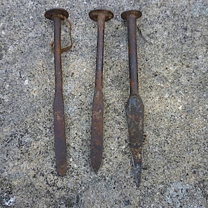 May include: Three antique iron spikes with a heavy rusted patina. Each spike features a flat, circular head and a long, straight shaft that tapers to a point. One spike includes a small, thin piece of twine looped through the head area.