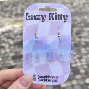 Peut inclure: Boucles d'oreilles en forme de nuage bleu clair avec des gouttes de pluie pendantes. Elles sont suspendues à des crochets argentés. Présentées sur une carte à carreaux lavande et blanche avec le texte "Lazy Kitty".