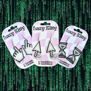 May include: Three pairs of earrings on pink and white cards, each with a different computer cursor design: a hand pointer, an arrow, and an hourglass. The cards are labelled "Lazy Kitty" with social media details.