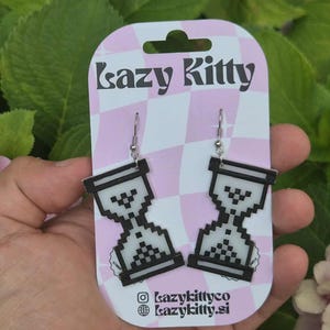 Könnte beinhalten: Pixelige Sanduhr-Ohrringe mit schwarzen Umrissen und weißer Füllung, hängend an silbernen Haken. Die Ohrringe werden auf einer rosa-weiß gemusterten Karte mit dem Text "Lazy Kitty" und Social-Media-Konten präsentiert.