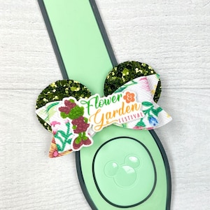 May include: A green and white floral bow with glitter accents and the text "Flower & Garden Festival" attached to a green Mickey Mouse ear headband.