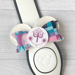 May include: A white Disney park MagicBand with a pink and blue patterned bow with a white button featuring the purple "Inside Out" logo. The bow is attached to the MagicBand with a white glitter circle.