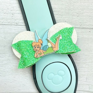 May include: Green glitter bow with a white glitter center featuring a Tinkerbell character illustration. The bow is attached to a light blue Mickey Mouse ear headband.