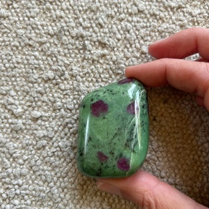 May include: A polished gemstone featuring a green base with dark green and red inclusions. The stone is held in a hand and has a smooth, rounded shape. The colors are natural and earthy, suggesting a mineral specimen.