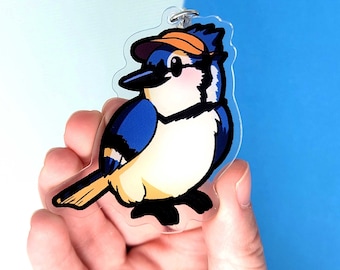 May include: A blue jay wearing an orange cap and sunglasses, with a white belly and black eyes. The bird is depicted in a cartoon style and is a charm on a clear acrylic keychain.