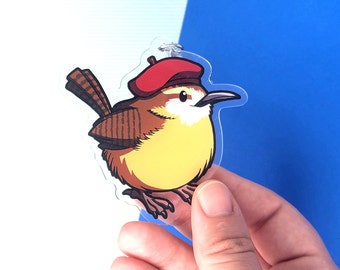 May include: A cartoon wren wearing a red beret, with brown and yellow feathers, is depicted on a clear acrylic charm.