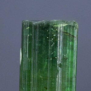 Op de afbeelding: Een levendig groene toermalijnkristal, verticaal geori&euml;nteerd, met een gestreepte textuur. De edelsteen is gemonteerd op een kleine, ruwe, grijze basis. De kleur van het kristal varieert van licht- tot diepgroen, met zichtbare interne structuren.