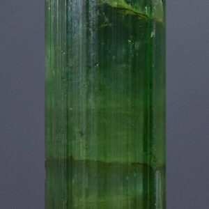 Op de afbeelding: Een doorschijnend, groene toermalijnkristal met verticale strepen. De edelsteen heeft een levendige, smaragdgroene kleur, met een enigszins ruwe textuur aan de bovenkant en een kleine, grijze basis. Het kristal is ongeveer 3,8 cm hoog.