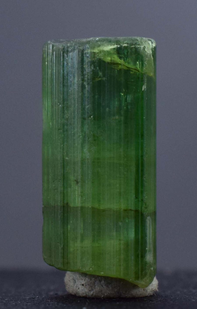 Op de afbeelding: Een doorschijnend, groene toermalijnkristal met verticale strepen. De edelsteen heeft een levendige, smaragdgroene kleur, met een enigszins ruwe textuur aan de bovenkant en een kleine, grijze basis. Het kristal is ongeveer 3,8 cm hoog.
