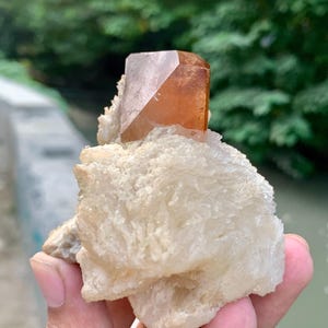 May include: A natural gemstone specimen featuring a large, amber-colored crystal with a geometric shape, set atop a cluster of white, textured mineral formations. The crystal is held in a hand, with a blurred green background.
