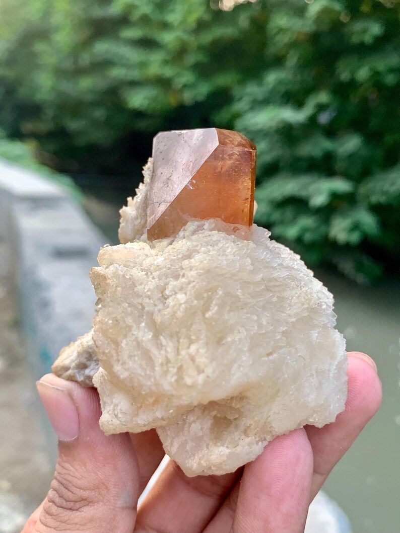 May include: A natural gemstone specimen featuring a large, amber-colored crystal with a geometric shape, set atop a cluster of white, textured mineral formations. The crystal is held in a hand, with a blurred green background.